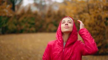 Kız yağmurun keyfini çıkarmak için kırmızı ceketini çıkarıyor, şehir parkında şemsiyesini açıp gökyüzüne bakıyor, yüzüne düşen yağmur damlalarının keyfini çıkarıyor, gülümsüyor, taze, mevsimsel iklim değişikliğinin tadını çıkarıyor.