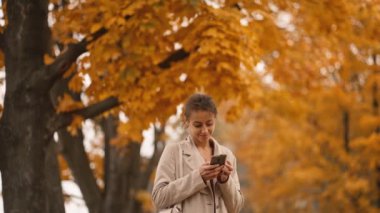 Sonbaharda parkta internette akıllı telefon kullanan mutlu, genç bir kadın. Çekici, sevimli, neşeli kadın dışarıda internet teknolojilerini kullanıyor. Akıllı telefon kavramı.