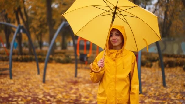 黄色いレインコートの女性と傘を持った女性は 子供のためのアトラクションと市立公園の雨の秋の日に散歩します 濡れた黄色いメープルは地面に葉 空っぽのベンチと遊び場でスイングします 落ちる気分 — ストック動画