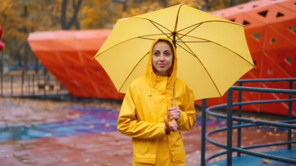 黄色いレインコートの女性と傘を持った女性は 子供のためのアトラクションと市立公園の雨の秋の日に散歩します 濡れた黄色いメープルは地面に葉 空っぽのベンチと遊び場でスイングします 落ちる気分 — ストック動画