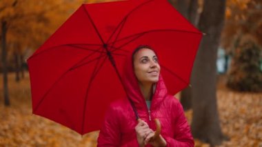 Yağmurlu bir günde, turuncu şehir parkında kırmızı şemsiyenin altında saklanan bir kadın yağmurun altında tek başına yürüyor. Yavaş çekim. Sonbahar sezonu ve şehir yaşam tarzı