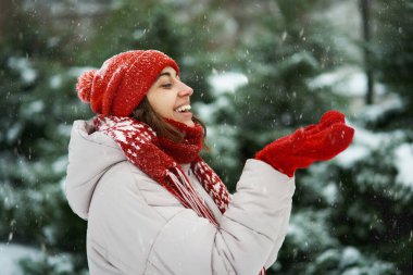 Kar parkında kırmızı şapkalı ve eşarplı mutlu bir kadın eldivenleriyle kar taneleri yakalıyor. Mükemmel mevsimlik an.