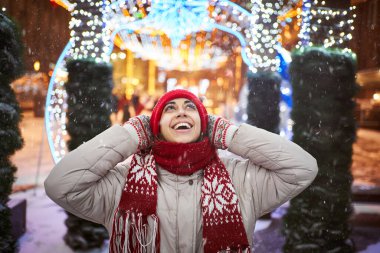 Kırmızı bereli ve eşarplı mutlu bir kadın yeni yıl gecesinde rengarenk ışıklarla şehir meydanında yürüyor.