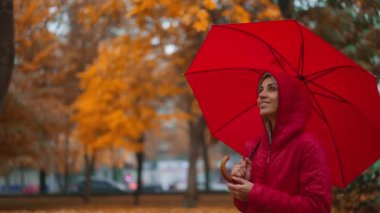 Yağmurlu bir günde, turuncu şehir parkında kırmızı şemsiyenin altında saklanan bir kadın yağmurun altında tek başına yürüyor. Yavaş çekim. Sonbahar sezonu ve şehir yaşam tarzı