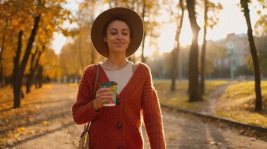 Sonbahar şehir parkında günbatımı güneşine karşı sarı yapraklı genç ve şık bir kadın. Şapkalı çekici bir kız, elinde bir fincan kahveyle sıcak bir sonbahar gününde yol boyunca parkta yürüyor..
