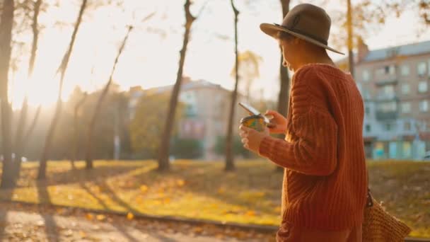 秋の公園の若いブルネットの女性の肖像画は 日没の太陽に対して黄色い葉を描いています 暖かい秋の日に公園でコーヒーと電話の紙コップの帽子でスタイリッシュな魅力的な女の子 — ストック動画