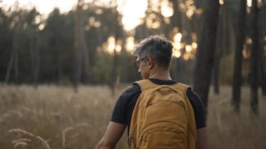 Yavaş çekim yetişkin gri saçlı, gözlüklü, sarı sırt çantalı adam sonbahar ormanında yol kenarında yürüyor. Sırt çantalı erkek yürüyüşçü yüksek otlarla ağaçların arasından yukarı çıkıyor. Seçici odaklanma. Ukrayna 'da seyahat