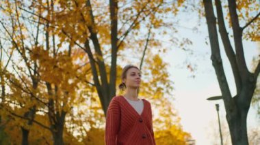 Sonbahar ormanında sarı yapraklı kırmızı örgü süveter giyen esmer kadın. Sıcak bir sonbahar gününde şık ve çekici bir kız parkta yürür. Yavaş çekim 4k görüntü