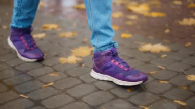 Spor ayakkabılı kadın bacakları, spor ayakkabıları. Yağmurlu sonbahar gününde parkta yürüyen bir kadın. Sarı akçaağaç yapraklı su birikintisine düşen yağmur damlaları şehrin kaldırımlarına yansıyor..