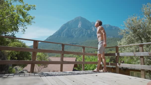 雄大な山を見下ろす景色のベランダで暖かい晴れた日を楽しむ運動体の大人のシャツレス男性 — ストック動画