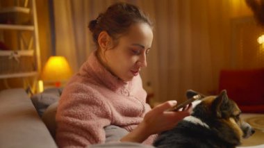 Evde köpeğiyle rahatlayan ve telefon kullanan genç bir kadının portresi. Şirin Galli Corgi köpeği sahibinin yanında dinleniyor, evde birlikte vakit geçiriyorlar. Dostluk köpekleri ve insanlar, sevimli anlar.