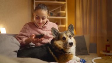 Evde köpeğiyle rahatlayan ve telefon kullanan genç bir kadının portresi. Şirin Galli Corgi köpeği sahibinin yanında dinleniyor, evde birlikte vakit geçiriyorlar. Dostluk köpekleri ve insanlar, sevimli anlar.