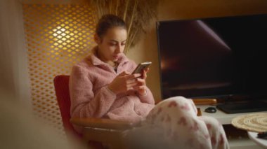 Evde takılan genç bir kadın evinde akıllı telefon cihazı kullanarak cep telefonu taşıyor. Gülümseyen kız blogcu yeni sosyal medya aboneliği, internetten satın alma, uygulamalarla ürünler sipariş etme.