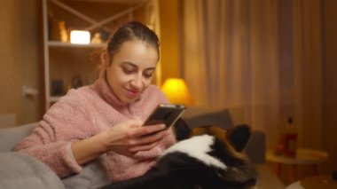Evde köpeğiyle rahatlayan ve telefon kullanan genç bir kadının portresi. Şirin Galli Corgi köpeği sahibinin yanında dinleniyor, evde birlikte vakit geçiriyorlar. Dostluk köpekleri ve insanlar, sevimli anlar.