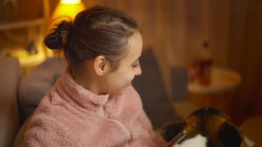 Evde köpeğiyle rahatlayan ve telefon kullanan genç bir kadının portresi. Şirin Galli Corgi köpeği sahibinin yanında dinleniyor, evde birlikte vakit geçiriyorlar. Dostluk köpekleri ve insanlar, sevimli anlar.