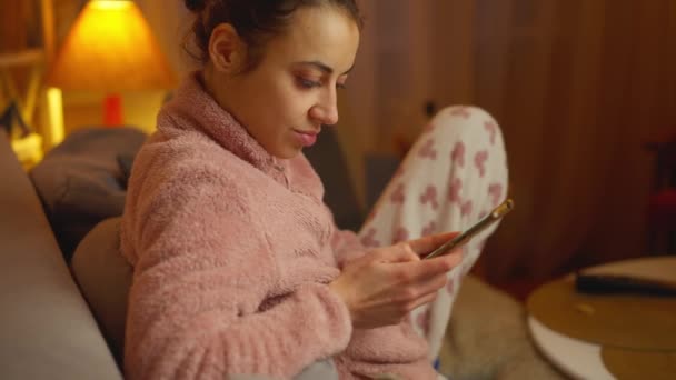 迷人的高加索女人穿着可爱的保暖休闲服 坐在舒适的客厅里的沙发上 带着电话 阅读新闻 浏览社交网络 网上订购 — 图库视频影像