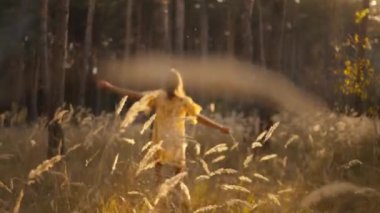 Sarı elbiseli genç esmer kadın gün batımında ormanda neşeyle koşuyor. Parkta uzun otlar ve harika sinema ışıklarıyla yürüyen mutlu bir kız. seçici odak, bulanık sahne