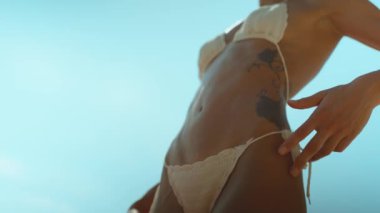 Sahilde gülümseyen bir kadının bikinisine ve tenine nazikçe dokunuşunun alçak açılı görüntüsü. Güneşli sahilde dinleniyor.
