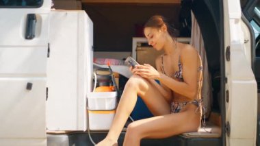 Bikinili gülümseyen kadın kampta oturuyor, akıllı telefon kullanıyor, daktilo çekiyor, kaydırıyor, sosyal medyayı sallıyor.