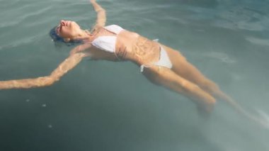 Kaygısız kadın, beyaz bikinili sakin sularda sırtüstü yüzüyor, huzur ve rahatlık saçıyor.