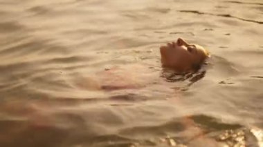 Kaygısız bir kadın, altın gün batımında huzur ve rahatlık içinde sırtüstü yüzüyor.