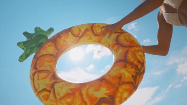 ralenti heureuse femme joyeuse en bikini blanc avec peau bronzée sur fond bleu ciel vomit grand anneau d'ananas flottant. fille profitant d'une chaude journée d'été ensoleillée en plein air. concept d'heure d'été.