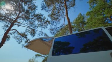 Kamp arabasının alt manzarası yeşil çam ağaçlarının ve mavi gökyüzünün arka planının kapıları açık. Açık havada seyahat, minibüs hayatı konsepti. Ormandaki araba. Yolculuk tutkusu