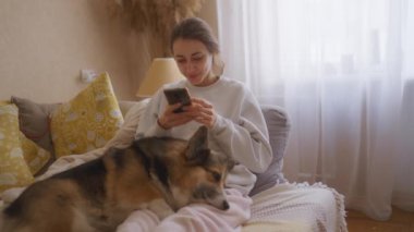 Genç bir kadın rahat bir kanepede uzanmış, akıllı telefonuyla sosyal medyada gezinirken Galli Corgi 'si bacaklarının üzerinde huzur içinde uyuyor.