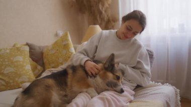 Kadın kanepeye uzanır ve Corgi bacaklarının üzerinde güzel bir şekilde uzanır. Köpeği nazikçe okşar, ona sarılır ve sevgiyle konuşur. Huzurlu ev ortamında evcil hayvan ve ev sahibi arasındaki bağı yakalayan iç açıcı sahne.