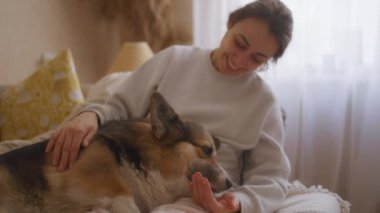 Kadın kanepeye uzanmış, Corgi bacaklarının üzerinde dinleniyor. Kıllı arkadaşını sımsıkı okşayıp kucaklarken köpek ellerini yalıyor. Sıcak atmosfer, aralarında paylaşılan sevgi ve ilgiyi vurguluyor.