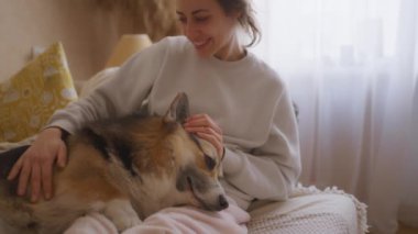 Rahat kadın, Corgi 'nin bacaklarına sarılıp rahat bir kanepeye uzanır. Köpeği sevgiyle okşar, evcil hayvan huzur içinde dinlenirken onunla nazikçe konuşur. Zaman sıcaklığı, özeni ve refakatin neşesini yayar..