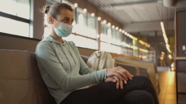 Tıbbi maskeli genç bir kadın klinik lobisinde bekliyor, sağlığı için endişeleniyor. Ayarlar tıp, güvenlik ve bulaşıcı hastalıkların önlenmesi temalarını yansıtıyor.