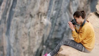 Uçurumun kenarında oturan kadın fotoğrafçı kameraya bakıyor ve fotoğraf çekiyor. Doğada çalışan seyahat ve tırmanma fotoğrafçıları. Kadın iş yerinde, yakalıyor.
