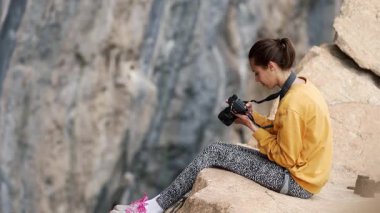 Uçurumun kenarında oturan kadın fotoğrafçı kameraya bakıyor ve fotoğraf çekiyor. Doğada çalışan seyahat ve tırmanma fotoğrafçıları. Kadın iş yerinde, yakalıyor.