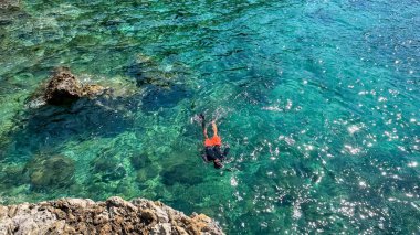 Budva, Karadağ - 18 Mayıs 2025: Erkek yüzücü kayalık kıyı şeridi yakınlarında güneş ışığıyla berrak turkuaz suya sahip