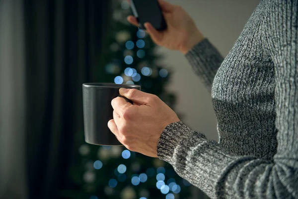 A young woman drinks a cup of tea or hot chocolate to warm up from the cold of winter. In the background the Christmas tree lights create a bokeh effect to celebrate the holy festivities.