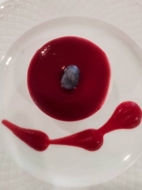 Yuvarlak tatlı kreması kırmızı böğürtlen sosuyla kaplı, üstünde yaban mersini olan, dekoratif soslu beyaz bir tabakta servis edilen.