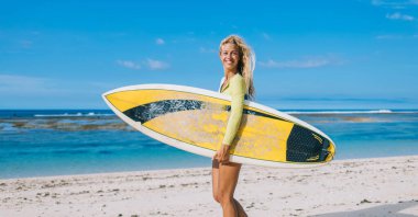Neşe dolu genç sörfçülerin yan görüntüsü kameraya bakıp tatilin tadını çıkarırken deniz kıyısında sörf tahtası ile durması.