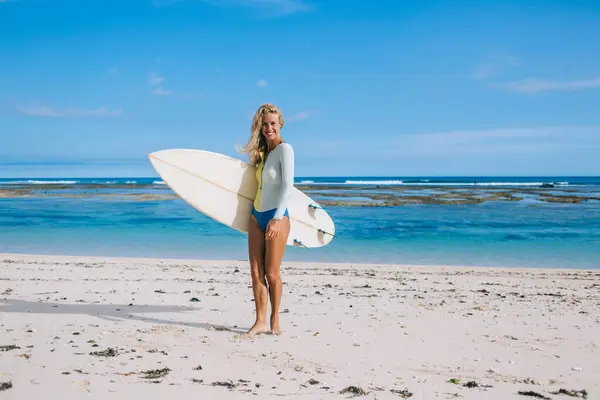 Joyful young attractive tanned woman with blond hair looking at camera while holding surf board and standing on resort beach