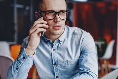 Gözlüklü dalgın genç adam akıllı telefondan arıyor ve ofiste bilgisayarla masada oturuyor. Netbook 'taki çalışma sürecinde cep telefonuyla iletişim kuran öğrenci.