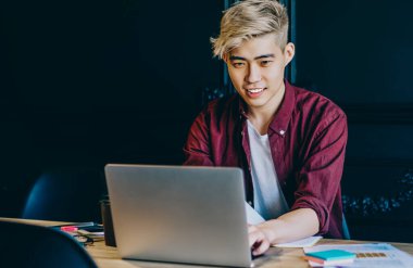 Pozitif genç adam Çin dizüstü bilgisayarında kablosuz internet bağlantısı kullanarak yazılımı güncelliyor. Günlük giyinmiş neşeli Japon hipster öğrenci internette komik webinar izlerken gülüyor.