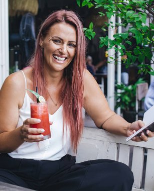 Smoothie ve kitapla dolu güzel, uzun saçlı kadın yaz günü kameraya gülümsüyor.