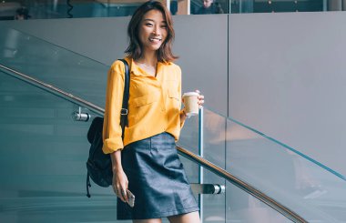 Tam vücut genç, pozitif, güzel, neşeli Asyalı, sarı tişörtlü, siyah deri etekli, sırt çantalı, Hong Kong 'da kahve ve cep telefonuyla merdivenlerden inen bir kadın.