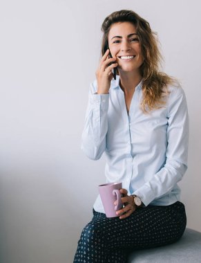 Genç, güzel bir bayan gülümsüyor ve bacak bacak üstüne atmış vaziyette oturuyor. Ofiste beyaz arka plan karşısında kameraya bakıyor.