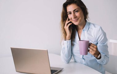 Genç esmer, gömlekli, beyaz masada dizüstü bilgisayarla oturmuş telefonla konuşurken gülümsüyor ve ofisteki kameraya bakıyor.