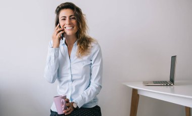 Genç, güzel bir bayan gülümsüyor ve bacak bacak üstüne atmış vaziyette oturuyor. Ofiste beyaz arka plan karşısında kameraya bakıyor.