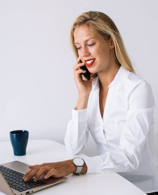 Masada oturan genç bayan çalışanın yan görüntüsü ve dizüstü bilgisayarda yazarken gülümsemesi ve ofiste beyaz arka planda akıllı telefonla konuşması.