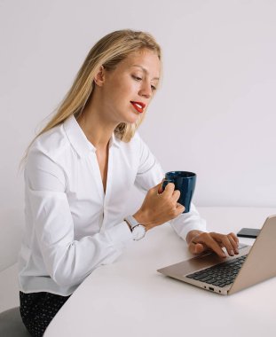 Masasında oturup dizüstü bilgisayarda beyaz arka planda yazı yazan, bardakta içkili genç bir kadının yan görüntüsü.