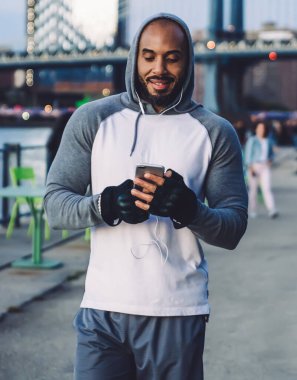 Gülümseyen atletik Afro-Amerikalı adam, rahat spor kıyafetleri içinde. Akşamları Megapolis 'teki rıhtımda dinlenirken akıllı telefondan mesaj atıyor.