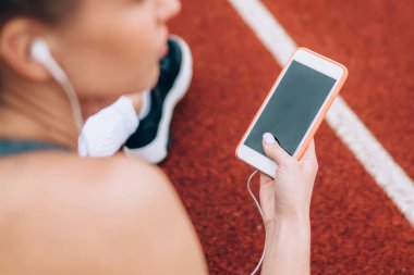 Arka planda, kulaklıklı, koşan pistte oturan ve müzik dinlemek için siyah ekranlı akıllı telefon kullanan belirsiz bayan sporcu görülüyor.